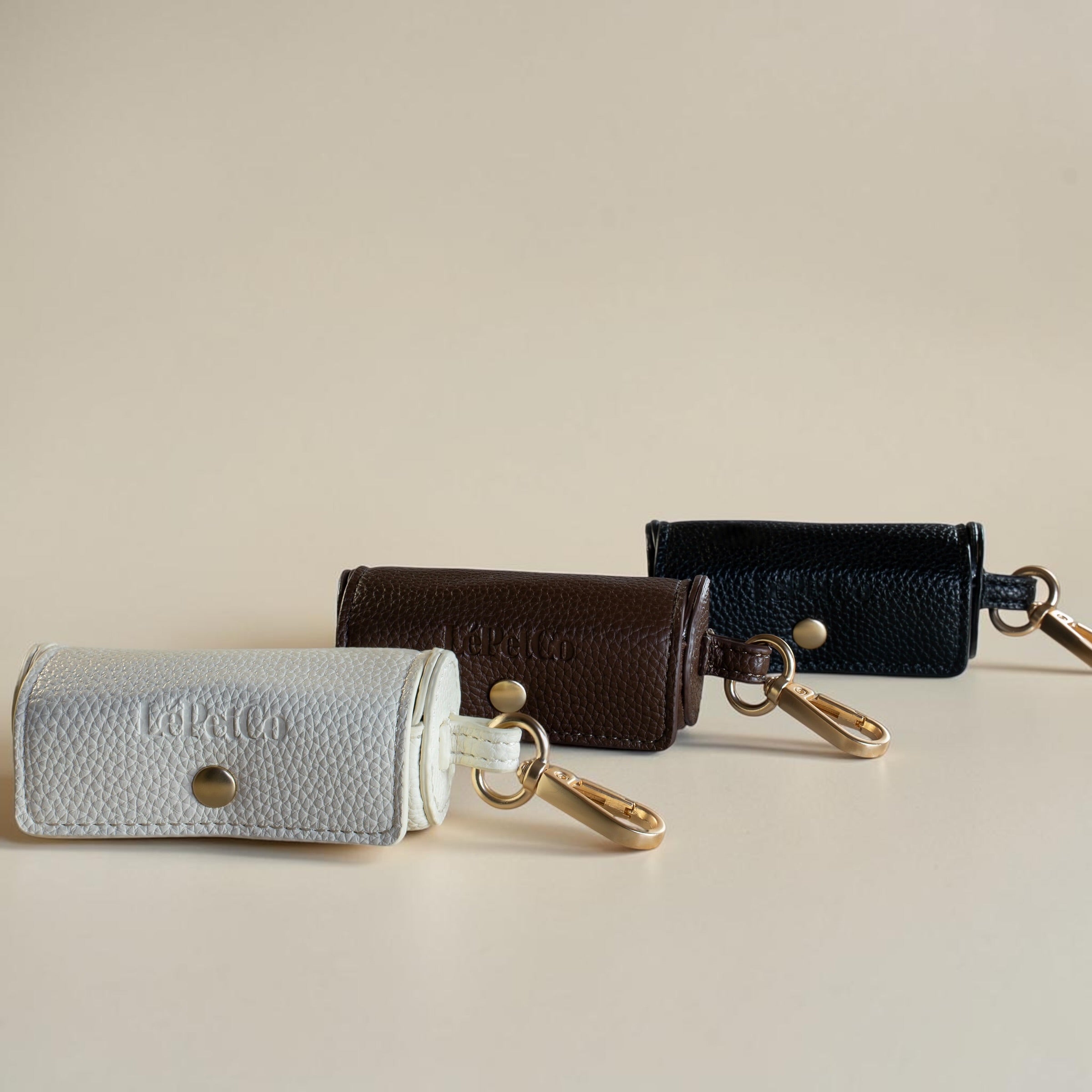 Three leather poop bag dispensers in white, brown, and black on a beige background